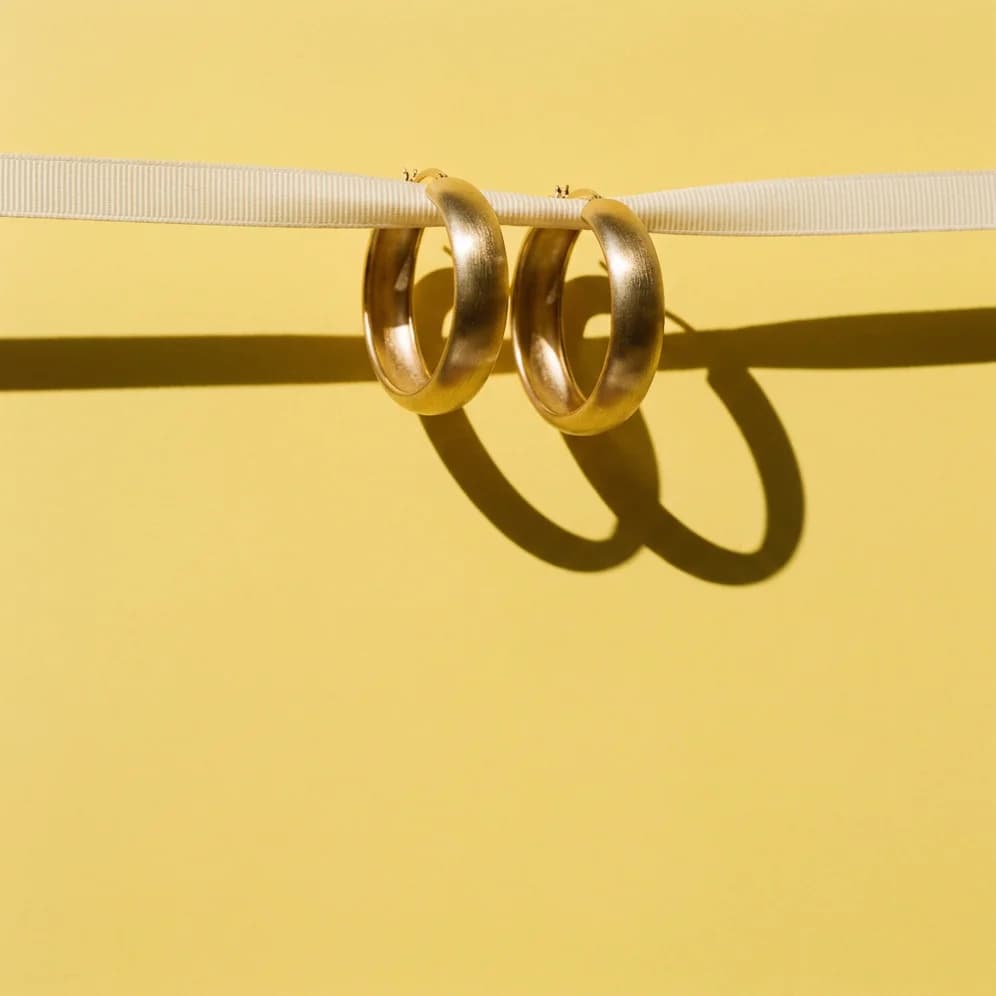Two mismatched gold hoop earrings on a bold butter-yellow field with graphic crossed shadows